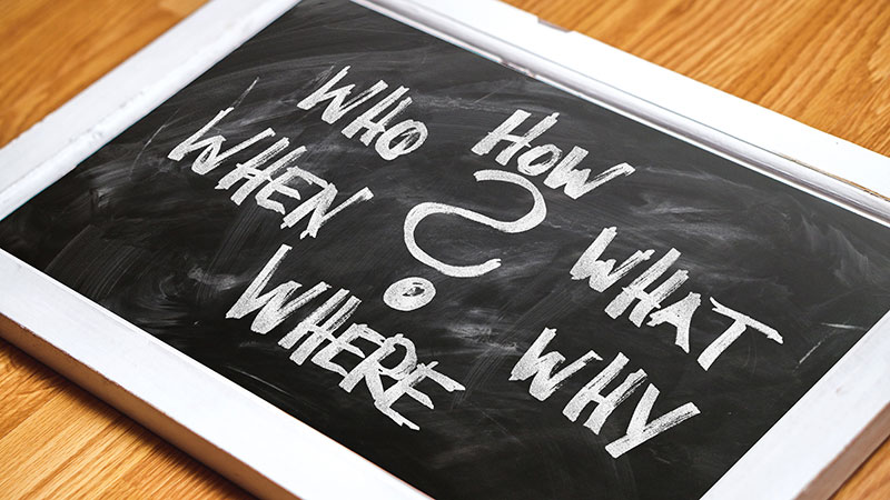 Small white framed chalk board laying on a table with a question mark in the center surrounded by the words; How, What, Why, Where, When, and Who.